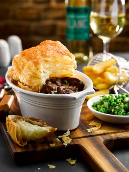Food photograph of a beef pie with a large crisp puff pastry lid, on a wooden chopping board with french fries and minted peas, with a glass of white wine in the background