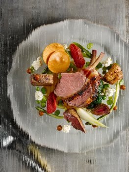 Aerial food photograph of a fine dining dish, juicy pink seared lamb cutlets with seared pigeon breast, spinach, asparagus, petit pois, feta cheese and dried plums served on a sheet of glass above a vintage white plate