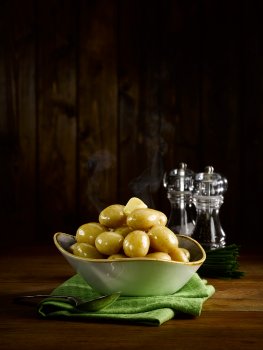 Food photograph of a bowl of steaming boiled baby potatoes, with a knob of butter slowly melting over them, served on a dark wood tabletop with a green napkin, and salt and pepper mills, and a dark wood background