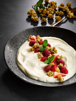 Food photograph of a vegan soya yogurt bowl topped with fresh raspberries, sliced fresh strawberries, sprigs of mint and crumbled granola  - served in a black ceramic bowl and shot on a dark set with scattered granola and mint leaves
