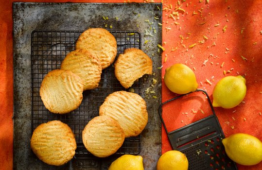 Aerial food photograph of lemon biscuits cooling on a wire rack, shot above a large vintage steel sheet alongside lemons and a vintage grater with scattered zest, on a bright red background