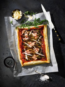 Aerial food photograph of a puff pastry tart topped with tomato sauce, grilled asparagus, rosettes of parma ham, parmesan shavings and drizzled with balsamic glaze, shot on a sheet of parchment paper on a black slate background