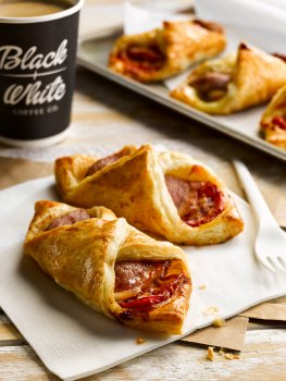 Food photograph of two crisp puff pastry turnovers filled with bacon and tomato, on a white napkin with a plastic fork, with three more turnovers and a takeaway cup of coffee in the background