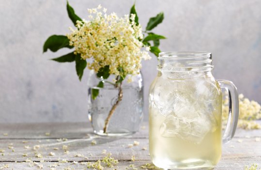 Drinks photograph of homemade elderflower cordial in a glass with ice, served with elderflowers in a glass jar, and elderflower petals scattered around the grey tabletop