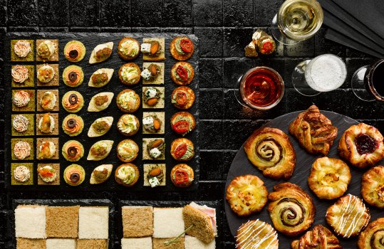Aerial food photograph of a sharing buffet spread, miniature sandwiches arranged into a chessboard pattern using white and brown bread, a selection of miniature viennoiserie, a selection of canapes and four glasses of prosecco, served on slate slabs on top of a black tiled tabletop