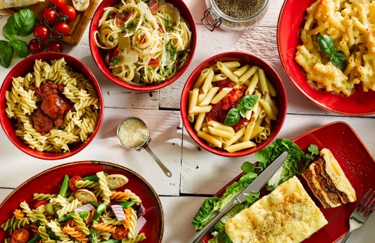 Aerial food photograph of a selection of pasta dishes served in red china bowls and plates, macaroni cheese, beef lasagne, penne arabbiata, spaghetti carbonara, fusilli with meatballs, and a tricolore fusilli pasta salad, garnished with vine cherry tomatoes, basil, parmesan, cracked black pepper and ciabatta