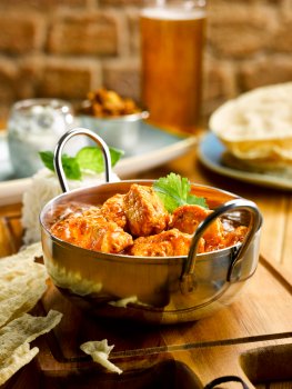 Food photograph, close up of a chicken tikka masala with thick shiny sauce in a silver balti dish served on a wooden board with shards of poppadom, and steamed white rice, served on a wooden table with a stack of poppadoms, a jar of raita and a ramekin of mango chutney, with a pint of lager