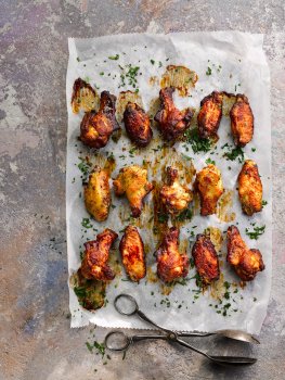 Aerial food photograph of baked chicken wings served on baking paper sprinkled with chopped parsley, wings glazed with different glistening sauces, alongside vintage serving tongs on a painted stone background