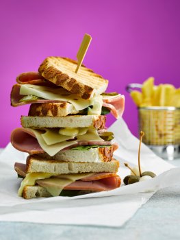 Close up food photograph of a toasted ham and cheese sandwich, thick slices of ham and melted swiss cheese between bar marked golden toasted tiger bread, served on baking paper with a basket of chips on a bright purple background