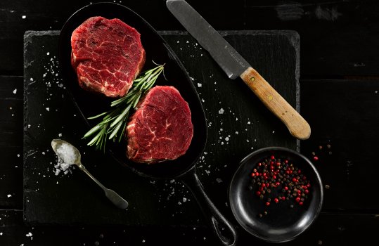 Aerial food photograph of a shiny raw beef fillet steak, presented in a cast iron pan on top of a slate board, garnished with sea salt and cracked black pepper, with a sprig of rosemary, served on a black wooden table with a small knife