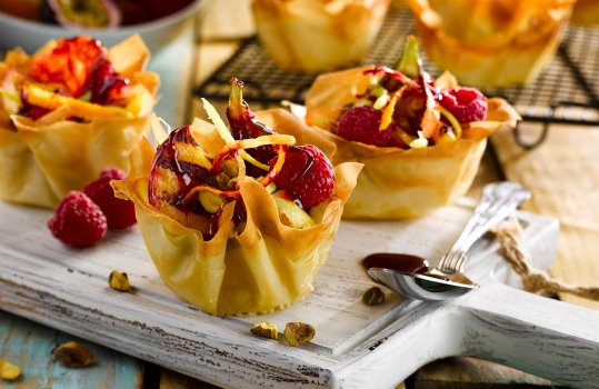 Food photograph of three grilled fruit cups made from crisp filo pastry, filled with grilled peaches, figs and raspberries; on a white chopping board garnished with pistachios