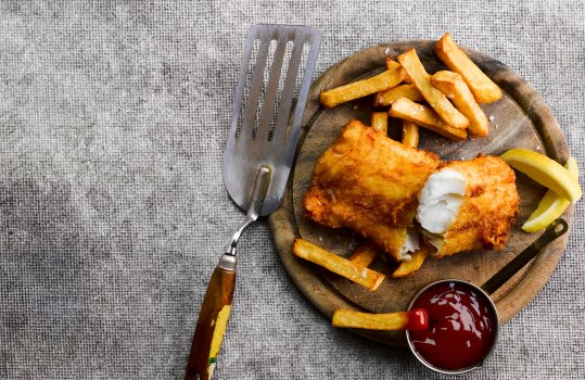 Aerial food photograph of homemade fish and chips, juicy flakey cod fillet encased in crisp golden batter, served on a wooden board with home made golden chips, lemon wedges, a pot of shiny tomato ketchup and a serving slice, on a grey canvas background
