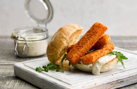 Food photograph of a fish finger sandwich, a toasted crispy bread roll with golden brown crisp fish fingers piled up on the bottom of the bun, served with tartare sauce and rocket leaves on a white board, on a grey tabletop with a grey wall in the background