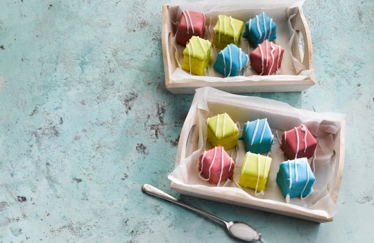 Food photograph of homemade fondant fancies, individual cubes of cake glazed with pastel coloured fondant and drizzled with white fondant, served on baking paper inside shallow wooden trays, on a sky blue background