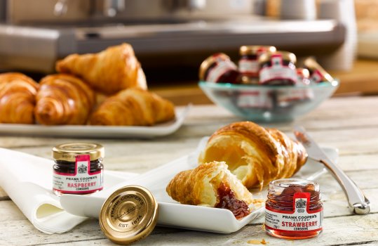 Food photograph of a continental breakfast, a crisp golden flakey croissant with strawberry jam served on a white wooden plate with individual pots of jam, a platter of croissants in the background with a bowl of pots of jam, and a coffee machine with cups in the background
