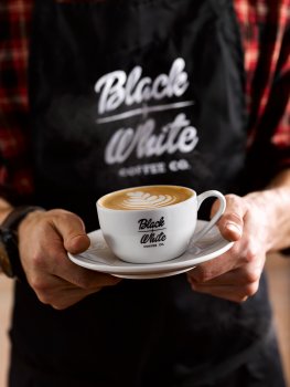 Drinks photograph of a barista in a branded apron holding a branded coffee cup and saucer, in the cup is a latte with a tulip made from the milk on the surface