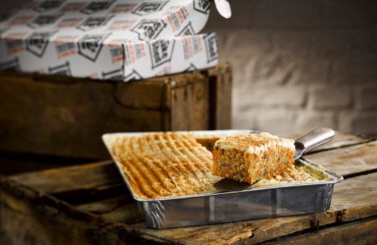 Food photograph of a spice dusted cream cheese icing topped carrot cake, a traybake in a foil tray with a slice removed and presented on a palette knife, served on a vintage apple crate with retail packaging in the background