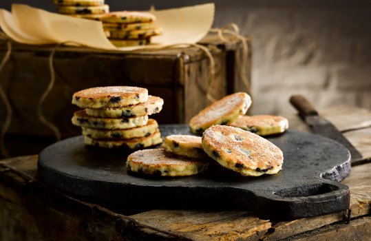 Food photograph of a selection of welsh cakes served on a traditional cast iron bakestone,  served on a vintage apple crate with retail packaging in the background