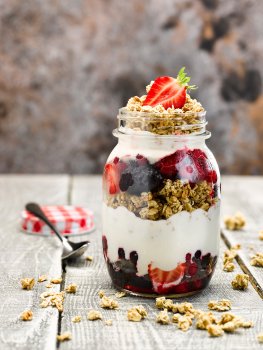 Food photograph of a layered granola, yogurt and summer fruit parfait, served in a jar with granola scattered around, on a bleached grey wooden tabletop and a dark background