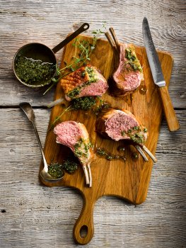 Aerial food photograph of a juicy pink rack of lamb with a crunchy seared exterior topped with basil and pine nut pesto, served on a wooden board garnished with thyme and presented on a grey wooden tabletop