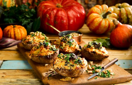 Food photograph of stuffed potato skins, potatoes baked and hollowed out before being filled up with cheesy mashed potato and baked again, served on a wooden board garnished with parsley and walnuts, presented on a wooden tabletop with a selection of autumnal squash in the background
