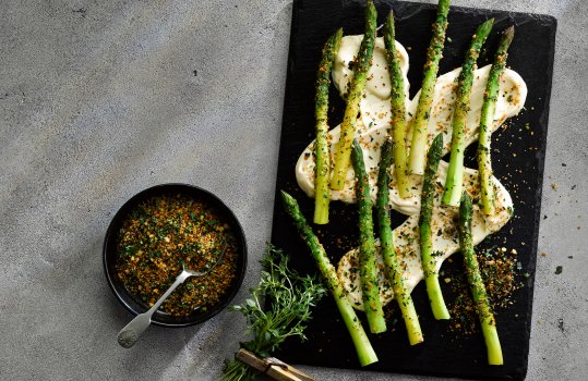 Aerial food photograph of steamed fresh asparagus spears served with cheese sauce and crispy herbed breadcrumbs, served on a slate slab on a grey concrete tabletop