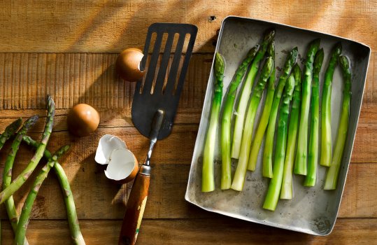 Aerial food photograph of fresh raw asparagus spears, peeled and placed into a tray of water, presented alongside raw unpeeled asparagus, eggshells and a spatula on a wooden table