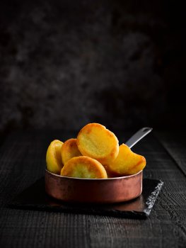 Food photograph of a small copper pan on a shiny slate filled with crisp golden roast potatoes, on a black wooden tabletop with a black background