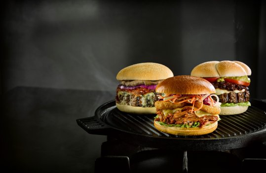 Food photograph of three topped burgers, a thick beef patty topped with blue cheese, red onion and a roasted mushroom, a pulled pork sandwich topped with cheese, onion rings and coleslaw, and a thick beef patty topped with cheese, salsa, sliced fresh tomato and guacamole. All three presented on a cast iron griddle with wisps of smoke on a dark grey background