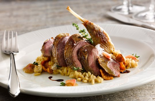 Food photograph of  a grouse and wild mushroom risotto, topped with a confit grouse leg and a juicy pink seared grouse breast, served on a white plate garnished with thyme sprigs on a grey wooden table