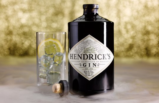 Drinks photograph of a bottle of Hendricks gin along with a glass full of ice and a slice of lemon, presented on a gold glittering background with clouds of smoke