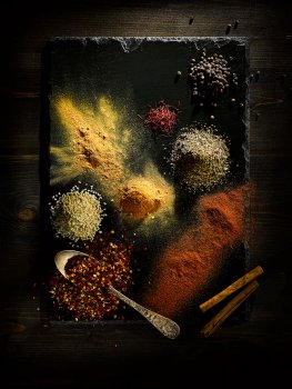 Aerial food photograph of a selection of dried spices, piles of dried chilli flake, sesame seeds, saffron, peppercorns, paprika, turmeric, mixed herbs, ginger and cinnamon sticks presented on a slate slab on top of a dark brown wooden table