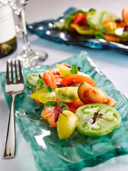 Food photograph of a heirloom tomato salad, green, yellow and red heirloom tomatoes with sea salt, basil and micro herbs, served on a teal glass plate on a white table, with a glass and bottle of white wine