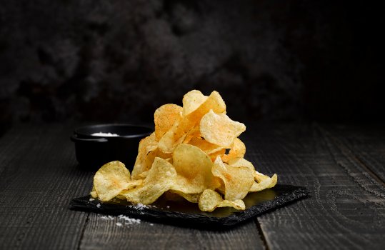 Food photograph of a pile of potato crisps on a shiny slate with a ramekin of sea salt, on a black wooden tabletop with a black background
