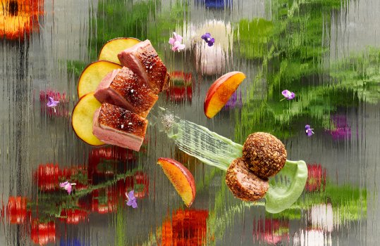 Aerial food photograph of a fine dining dish, seared crispy juicy pink duck breast served with grilled peach slices, a deep fried confit duck leg bonbon, served on a glass sheet above a selection of fruit and flowers