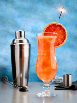 Food photograph of an orange and grapefruit mocktail served in a hurricane glass garnished with a slice of pink grapefruit and a lit sparkler, served on a grey tabletop with a cocktail shaker, a bar spoon and a spirit measure, with a bright blue background