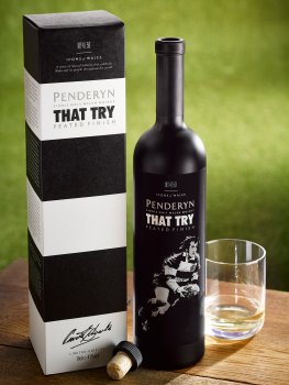 Drinks photograph of a limited edition Penderyn Welsh whisky, commemorating a rugby match between The Barbarians and the All Blacks, shot on a wooden table in an outdoor setting, with a glass of whisky next to the bottle and the retail packaging