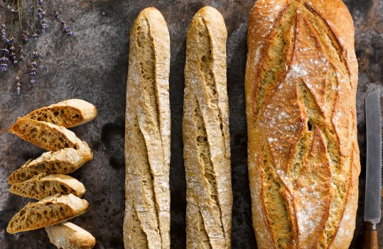 Aerial food photograph of artisan sourdough bread, a large loaf and two baguettes shot on a dark background with sliced toasted golden baguette, lavender sprigs and a vintage bread knife