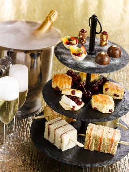 Food photograph of a festive afternoon tea featuring smoked salmon finger sandwiches, fruit scones with jam and clotted cream, and miniature Christmas puddings, alongside two glasses of champagne, and a champagne bottle in an ice bucket, all on a shiny gold background