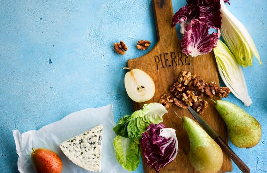 Aerial food photograph of ingredients to make a pear and roquefort salad, fresh juicy seasonal pears with fresh crunchy radicchio and white chicory, shot on a wooden board with walnut halves and roquefort cheese, on a bright blue background