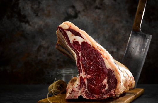 Food photograph close up of a glistening bone in dry aged rib of beef shot on a wooden board with butchers twine, sea salt and a vintage meat cleaver in the background