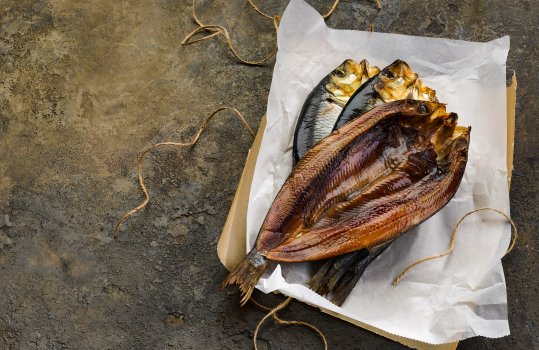 Food photograph close up of a stack of whole smoked kippers, whole butterflied glistening smoked fish with a dark mahogany flesh from being smoked, served on wax paper with twine on a brown stone background