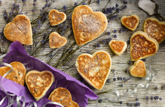 Aerial food photograph of homemade lavender Welsh cakes, heart shaped lavender flecked Welsh cakes served in a purple paper bag, along with sprigs of lavender on a bleached wooden background