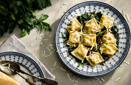 Food photograph of a bowl of agnolotti, rectangles of pasta filled with pureed potato, alongside crispy fried leeks, with parsley and parmesan; on a grey background