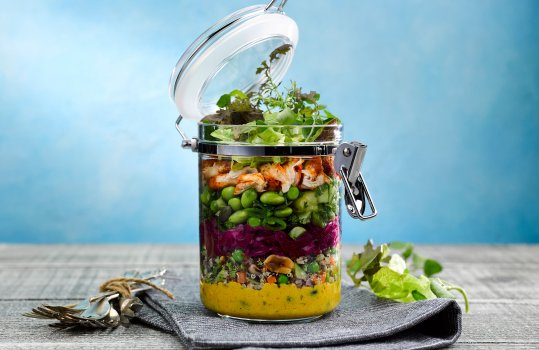 Food photograph of a layered salad in a takeaway jar, mixed vegetables with three colour quinoa, pickled red cabbage, edamame beans, tikka spiced chicken and mixed leaves with micro herbs served in a plastic sealable jar, on a grey napkin with a bundle of vintage forks and salad leaves to garnish, on a light grey tabletop with a bright blue background