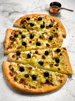 Food photograph of a homemade pissaladiere, a flatbread topped with caramelised onions and black olives, sliced into wedges and served on a marble background