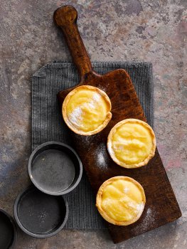 Aerial food photograph of homemade individual lemon tarts, crisp short pastry cases filled with rich homemade lemon custard and sprinkled with icing sugar, served on a wooden board with a grey napkin