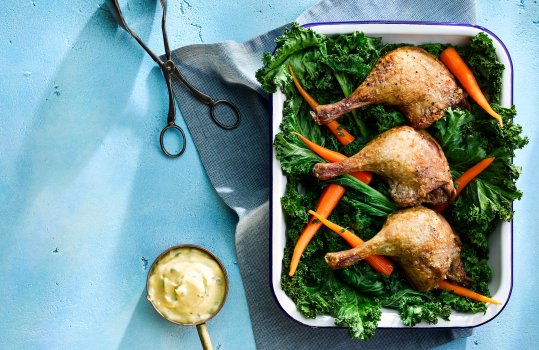 Aerial food photograph of slow cooked crispy roasted duck legs on a bed of kale and pickled carrots, served in an enamel dish with a copper pot of bernaise sauce on a bright blue background with vintage serving tongs