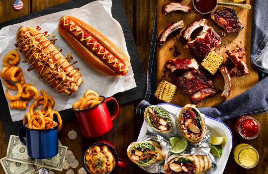 Food photograph of an Americana sharing table, featuring hot dogs in brioche buns with mustard, ketchup and crispy onions alongside curly fries and macaroni cheese served in enamel cups, a large wooden chopping board with shiny glazed barbecue pork ribs and bar marked grilled corn, and an enamel dish filled with chicken burritos, and lime wedges. All on a dark wooden tabletop with american flag pin badges, and dollar bills