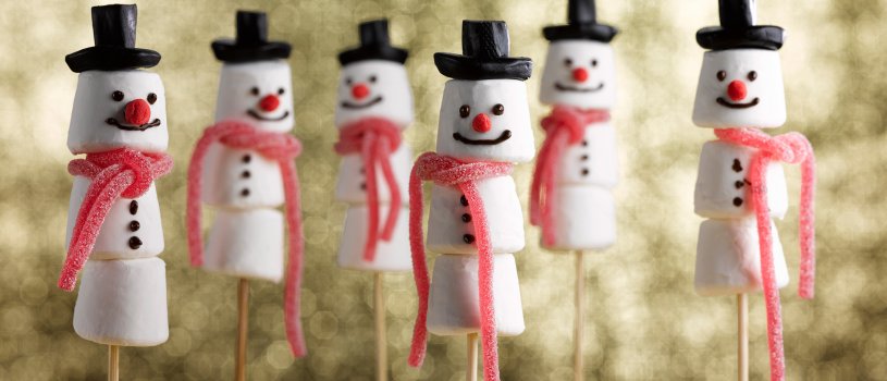 Food photograph close up of a group of marshmallow snowmen, marshmallows threaded onto skewers and decorated to look like snowmen, with liquorice hats and strawberry lace scarves, shot in a festive setting on a glittering gold background